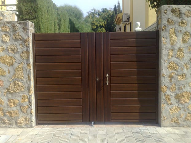 Puerta abatible de 2 hojas en lamas de aluminio extrusionado lacada madera. Disponible en varios colores y fabricada a medida.